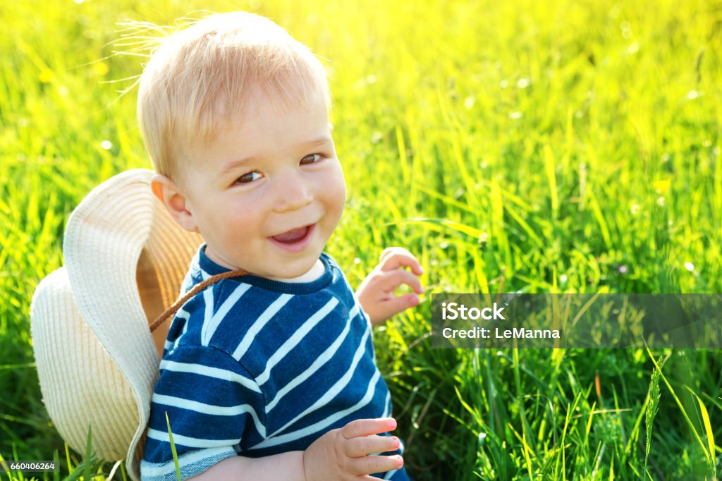 Happy baby boy sitting in grass on the fieald with dandelions at sunny summer evening. Smiling child outdoors